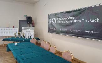 baner z napisem XXV Międzynarodowe Mistrzostwa Polski w Tarokach, stół, na drugim planie puchaty, upominki oraz tablica wyników